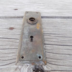 May include: A rectangular, weathered brass door plate featuring a central circular knob housing and a keyhole below. The metal surface displays a tarnished, aged patina with visible oxidation, four small screw holes, and beveled edges.