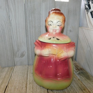 May include: A vintage ceramic cookie jar shaped like a woman wearing a pink dress and a green apron. The jar has a lid with a floral design.