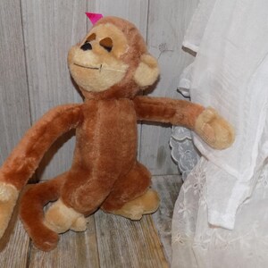 May include: A plush brown monkey with a light tan face, hands, and feet. The monkey has a small pink bow on its head and is posed with its arms outstretched. The toy is made of soft, fuzzy material.