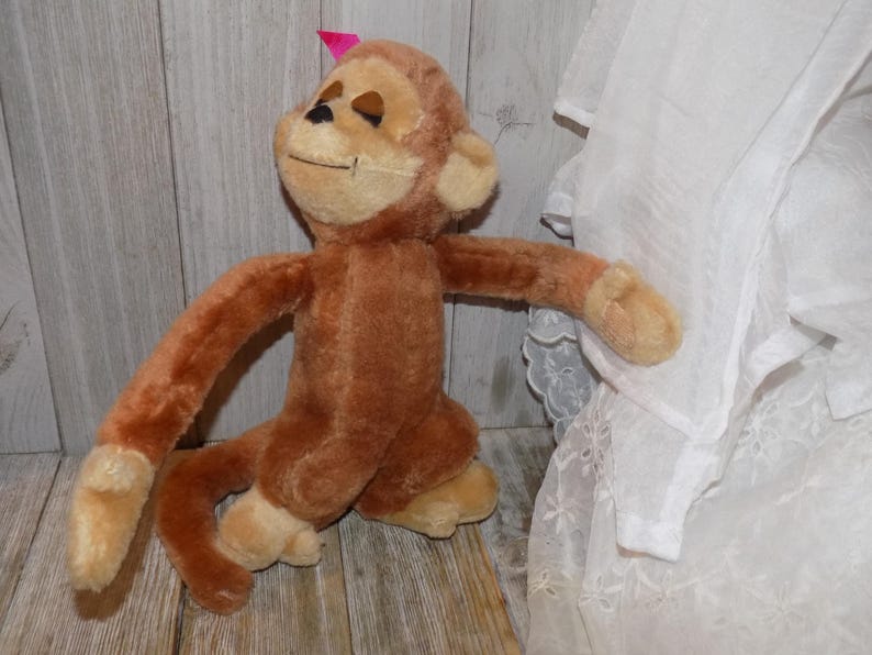 May include: A plush brown monkey with a light tan face, hands, and feet. The monkey has a small pink bow on its head and is posed with its arms outstretched. The toy is made of soft, fuzzy material.