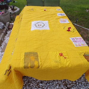 May include: A handmade yellow quilt with various embroidered and printed patches. The quilt features a grid pattern and includes images of a rattle, a baby, and text. The quilt is spread over a table outdoors.
