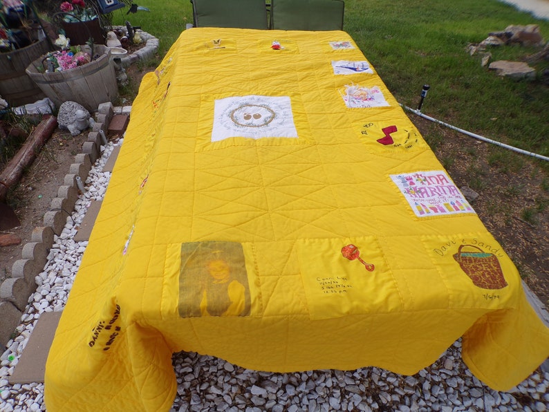 May include: A handmade yellow quilt with various embroidered and printed patches. The quilt features a grid pattern and includes images of a rattle, a baby, and text. The quilt is spread over a table outdoors.