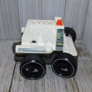 May include: A vintage toy vehicle in off-white and gray, featuring large black wheels and a detailed control panel. The toy has an orange light and various molded details, suggesting a retro design.