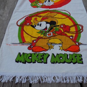 Peut inclure: Serviette en coton blanc avec une illustration color&eacute;e de Mickey Mouse en tenue de cow-boy, avec un lasso. La serviette a un bord &agrave; franges et les mots "Mickey Mouse" en vert.
