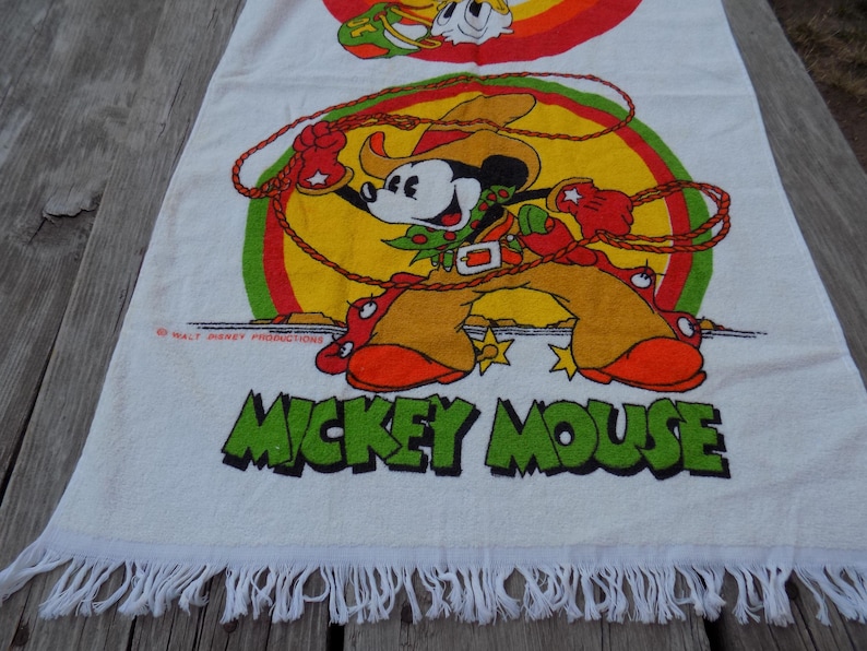 Peut inclure: Serviette en coton blanc avec une illustration color&eacute;e de Mickey Mouse en tenue de cow-boy, avec un lasso. La serviette a un bord &agrave; franges et les mots "Mickey Mouse" en vert.