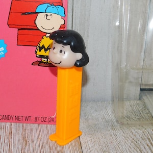 May include: A vintage orange Pez dispenser featuring a cartoon image of Lucy from the Peanuts comic strip. The dispenser is in its original packaging, which includes the text "Fun 'n Games On Back" and "TOTAL CANDY NET WT. .87 OZ. (24.7".