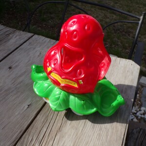 May include: A red plastic strawberry shaped toy with green leaves. The toy has the words "Strawberry Shortcake" printed on it.