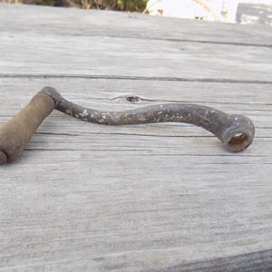 May include: A vintage metal hand crank with a weathered, dark grey finish and a cylindrical wooden handle. The crank features a curved arm design with visible signs of oxidation and wear, ending in a circular attachment point with a central hole.