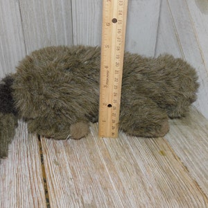 Puede incluir: Un juguete de peluche de mapache marr&oacute;n y negro acostado sobre una superficie de madera. El juguete mide aproximadamente 25 cm de largo y tiene una cola larga y tupida.