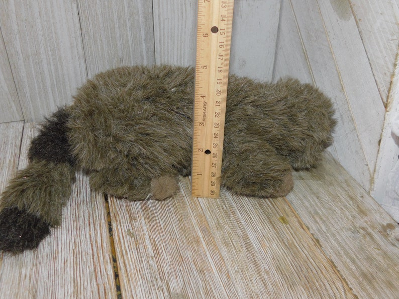 Puede incluir: Un juguete de peluche de mapache marr&oacute;n y negro acostado sobre una superficie de madera. El juguete mide aproximadamente 25 cm de largo y tiene una cola larga y tupida.