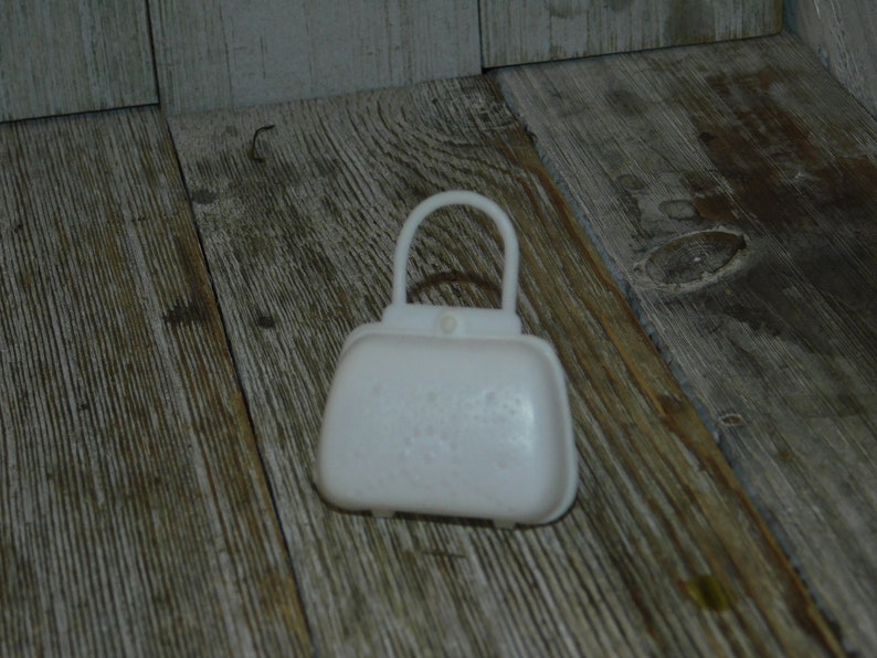 Op de afbeelding: Een kleine, witte handtas met een gebogen handvat. De tas heeft een rechthoekig lichaam met afgeronde randen en een klein knoopdetail. De achtergrond is een houten oppervlak.