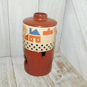May include: A brown ceramic jar with a geometric design of houses and trees in blue, red, and beige. The jar has a lid and a hole in the bottom.