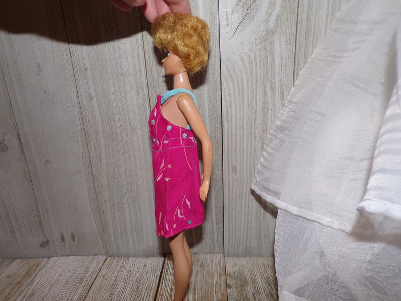 May include: A vintage Barbie doll wearing a pink dress with a floral pattern and a blue ribbon around her neck. The doll has blonde hair and blue eyes.