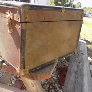 May include: Vintage brown trunk with brass hardware. The trunk has a rectangular shape with metal corner protectors and a latch closure. The trunk is resting on a wooden structure, suggesting it is an antique or decorative storage item.