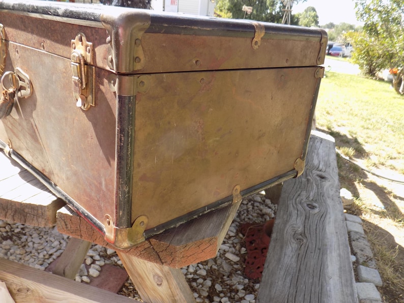 May include: Vintage brown trunk with brass hardware. The trunk has a rectangular shape with metal corner protectors and a latch closure. The trunk is resting on a wooden structure, suggesting it is an antique or decorative storage item.
