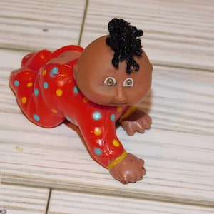 May include: A vintage crawling baby doll with a brown complexion, wearing a red onesie with blue and yellow polka dots. The doll has black hair and is posed on a wooden surface.