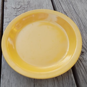 May include: A round, yellow plate with a slightly raised rim. The plate is a solid, warm yellow color and appears to be made of ceramic or a similar material. The plate is sitting on a weathered, gray wooden surface.