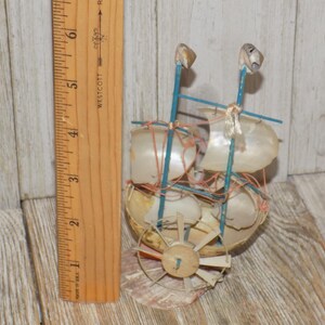May include: A handcrafted miniature sailboat made from seashells, with blue supports and white sails. The decorative nautical piece includes a wooden rudder and is approximately 4 inches tall.