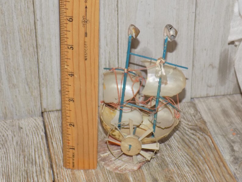 May include: A handcrafted miniature sailboat made from seashells, with blue supports and white sails. The decorative nautical piece includes a wooden rudder and is approximately 4 inches tall.