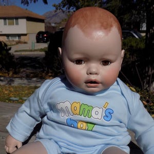 May include: A baby doll with a smooth, light-toned head and brown eyes, wearing a light blue long-sleeved shirt. The shirt features the text "mama's boy" printed in white, yellow, and blue letters across the chest area.