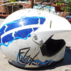 May include: White motorcycle helmet with a blue and white geometric design and the word "FGX" written in black. The helmet has a black visor and is slightly dirty.