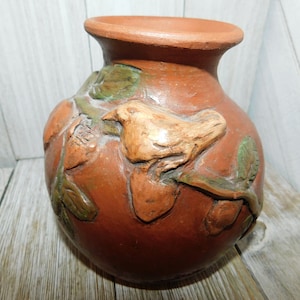 May include: A terracotta vase with a bird and leaves sculpted into the surface. The vase is a reddish-brown color with green and brown accents.