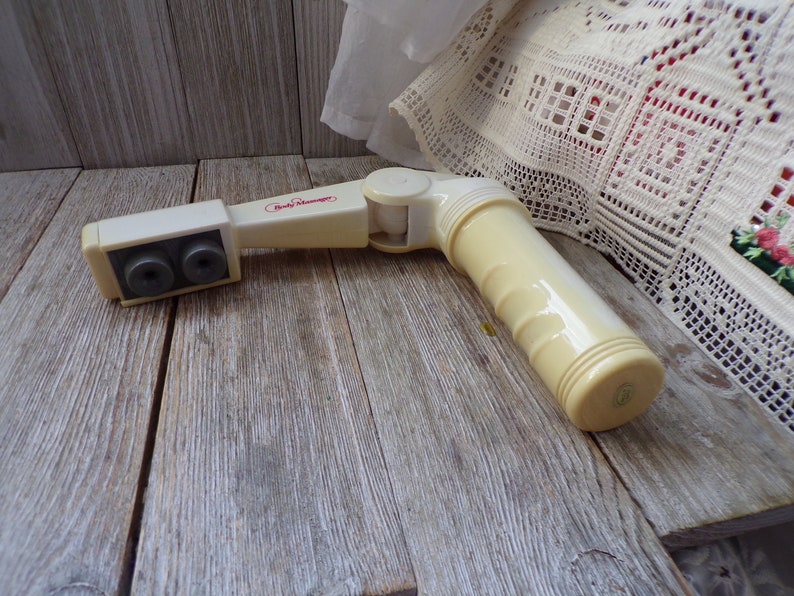 May include: A white plastic body massager with two round heads and a swivel joint. The massager has the words "Body Massager" printed on it in red.