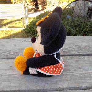 May include: A plush Minnie Mouse doll, seated on a wooden surface. The doll has black fur, a cream face, and orange accents on the feet. It wears a white apron with orange polka dots and a yellow bow.