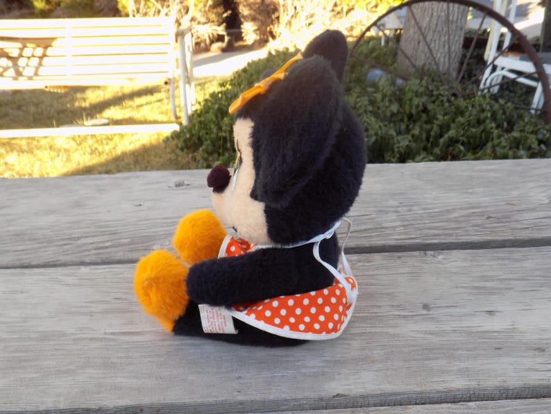 May include: A plush Minnie Mouse doll, seated on a wooden surface. The doll has black fur, a cream face, and orange accents on the feet. It wears a white apron with orange polka dots and a yellow bow.