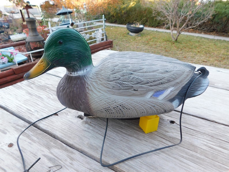 May include: A plastic decoy duck with a green head and a gray body. The duck is sitting on a yellow base and is attached to a black string.