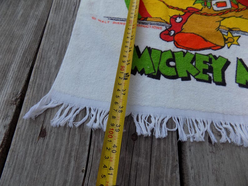 Peut inclure: Serviette en coton blanc avec un motif Mickey Mouse color&eacute; et le texte "MICKEY MOUSE". La serviette a une bordure &agrave; franges et mesure environ 104 cm de long.