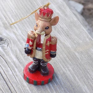 May include: A mouse figurine ornament wearing a red and gold military-style uniform with a matching crown. The mouse has brown fur, black eyes, and holds a red and white striped staff. It stands on a round red base with green stripes and a gold cord.