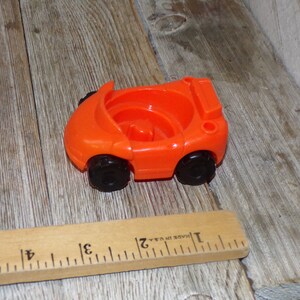 May include: Orange plastic toy car with black wheels. The car is approximately 5 cm long.