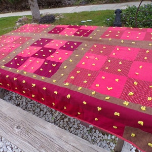 May include: A red and brown patchwork quilt with yellow knotted fabric accents. The quilt is laid out on a wooden table.