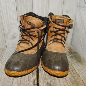 May include: A pair of brown leather and black rubber duck boots with a yellow trim. The boots have laces and the text "Thinsulate" is visible on the right boot.