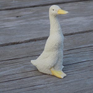 May include: A white goose figurine with a textured, snow-like surface finish. The goose has a long neck, a small black eye, and a smooth, pale yellow beak and webbed feet. The figure is depicted in a standing position, facing toward the right side.