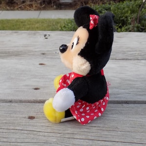 May include: A plush Minnie Mouse doll, featuring black ears, a red bow, and a red dress with white polka dots. The doll has a cream-colored face and yellow shoes. The toy is sitting on a weathered wooden surface.