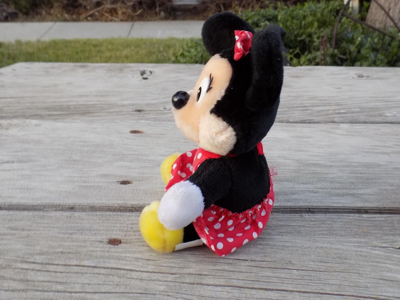 May include: A plush Minnie Mouse doll, featuring black ears, a red bow, and a red dress with white polka dots. The doll has a cream-colored face and yellow shoes. The toy is sitting on a weathered wooden surface.