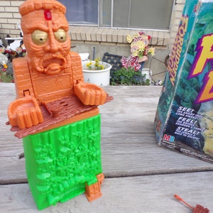 May include: A brown and green plastic game piece shaped like a head with a red jewel on the forehead. The game piece is part of the board game "Forbidden Island".