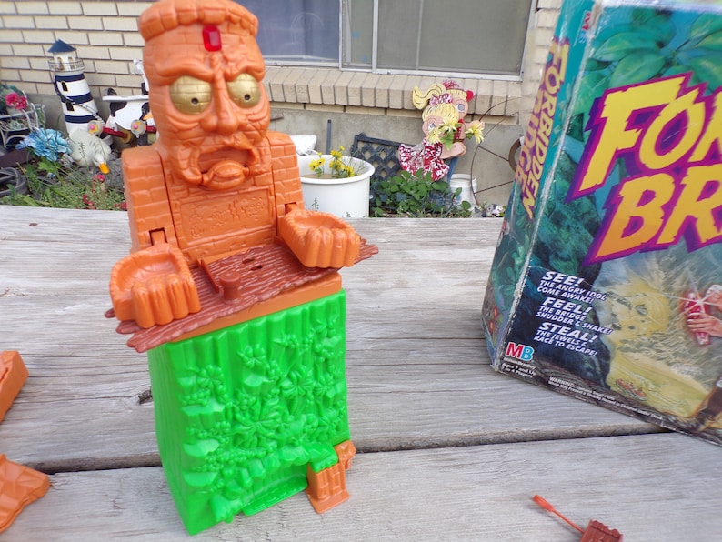 May include: A brown and green plastic game piece shaped like a head with a red jewel on the forehead. The game piece is part of the board game "Forbidden Island".