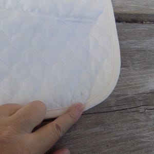 Op de afbeelding: Witte quilted stof met een witte binding rond de rand. De stof heeft een ruitpatroon.