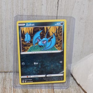 May include: A Pokémon trading card featuring Zubat, a blue bat-like creature with large wings. The card has a yellow border and the text "Zubat" at the top. The card is in a protective sleeve.