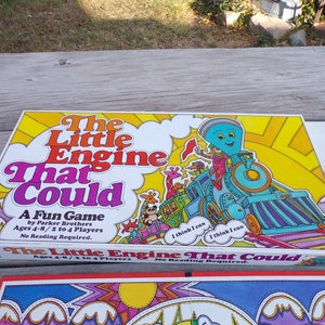May include: A vintage board game box featuring a colorful illustration of a blue train engine with a smiling face. The box is yellow with orange and red accents. The game is called "The Little Engine That Could" and is for ages 4-8 with 2 to 4 players. No reading is required.