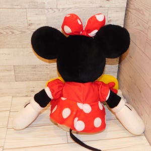 Op de afbeelding: Een pluche Minnie Mouse pop, van achteren gezien, met grote zwarte oren en een rode strik met witte stippen. Ze draagt een rode jurk met witte stippen en witte handschoenen. De pop heeft een zwarte staart.