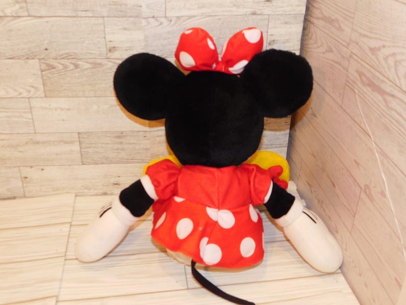 Op de afbeelding: Een pluche Minnie Mouse pop, van achteren gezien, met grote zwarte oren en een rode strik met witte stippen. Ze draagt een rode jurk met witte stippen en witte handschoenen. De pop heeft een zwarte staart.
