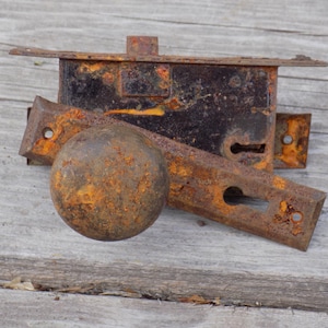 May include: An assortment of antique, rusty metal hardware components, including a door latch, a handle, and a strike plate. The pieces are heavily corroded with orange and brown rust, suggesting age and exposure to the elements. The items are arranged on a weathered wooden surface.
