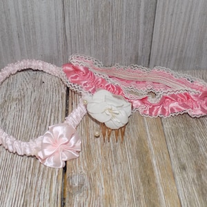 May include: A set of pink and white hair accessories, including a headband, a comb with a flower, and a piece of lace trim.