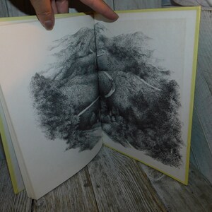 May include: An open book with a black and white illustration of a winding road through a mountainous landscape. The book has a yellow cover.