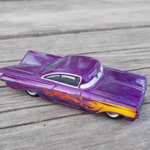 May include: A purple toy car with flame decals in orange and yellow. The car has a stylized design with large fins and a smiling face on the windshield. The toy car is resting on a wooden surface.