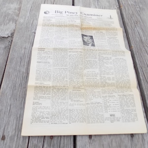 May include: A vintage copy of the Big Piney Examiner newspaper printed on off-white paper. The front page features multiple columns of black text, several headlines, and a small black and white portrait photograph of a person in the upper section.